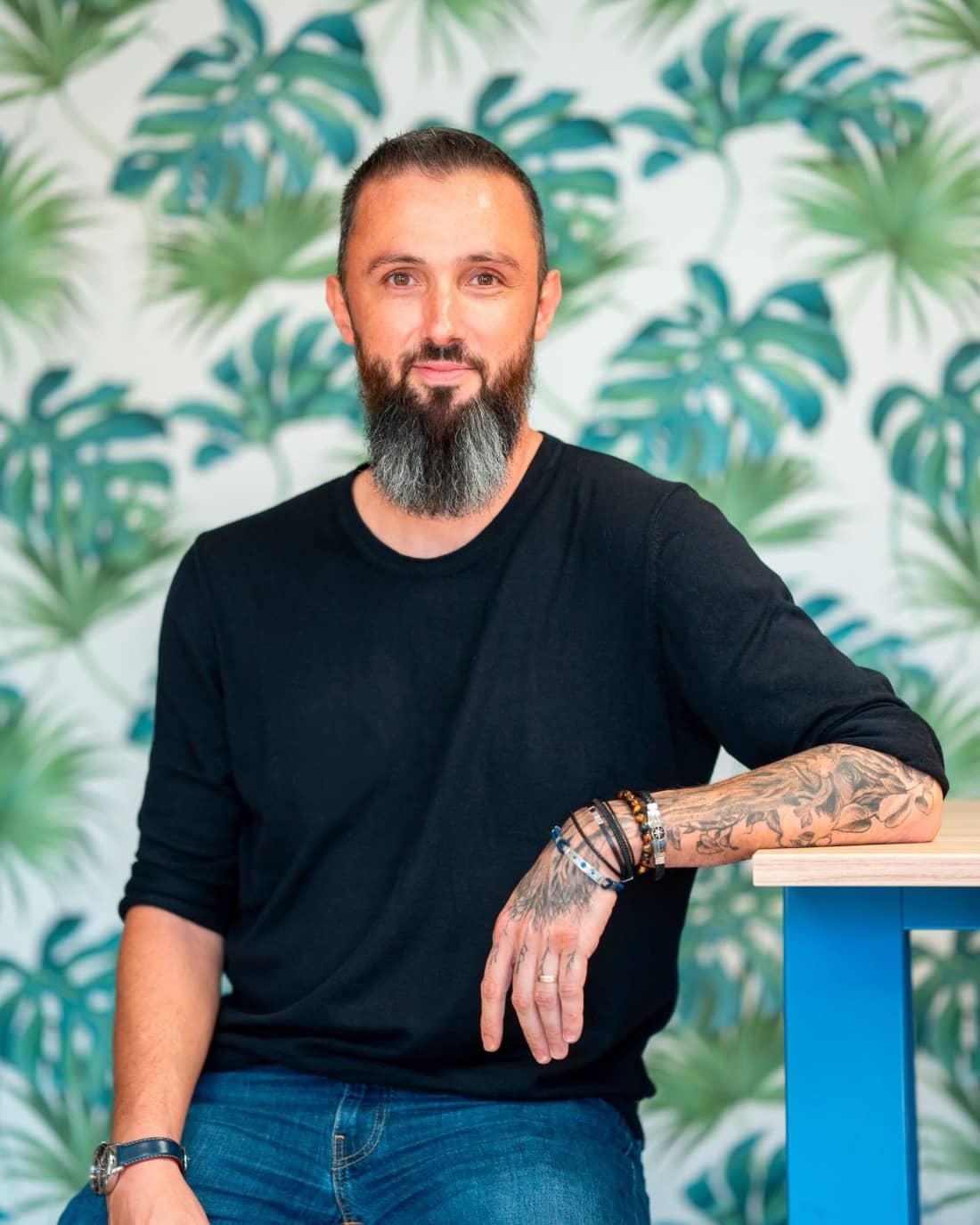 Souriant, assis, un avant-bras posé sur une table, en haut noir devant un papier peint à feuilles tropicales.